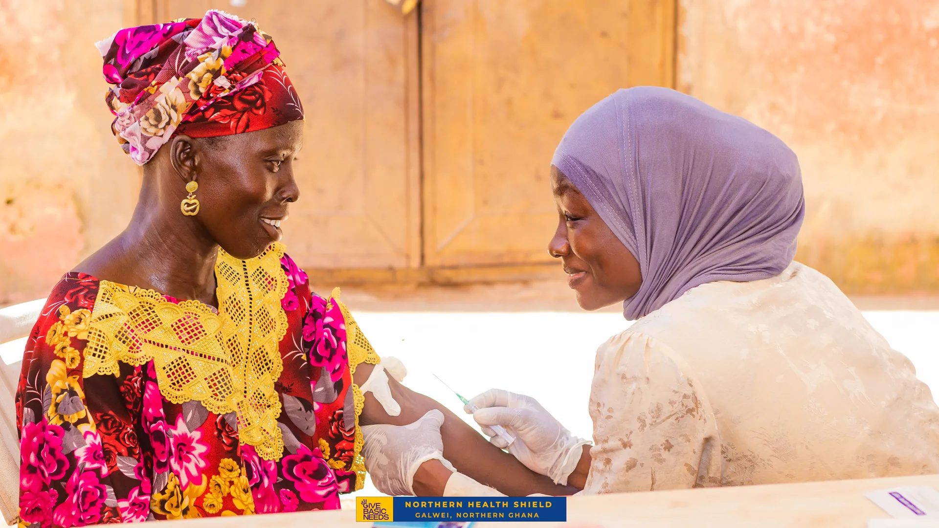 A smiling woman receives a vaccination from a Northern Health Shield health worker in Galwei, Northern Ghana